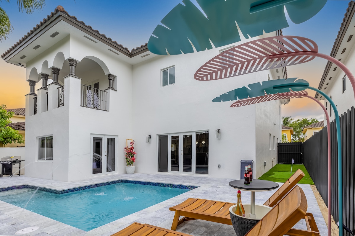 A modern exterior of the house showcases a private pool with a surrounding patio featuring loungers. Tall, elegant arches and windows allow for ample natural light, while tropical leaves in the foreground add a vibrant touch to the inviting outdoor space.