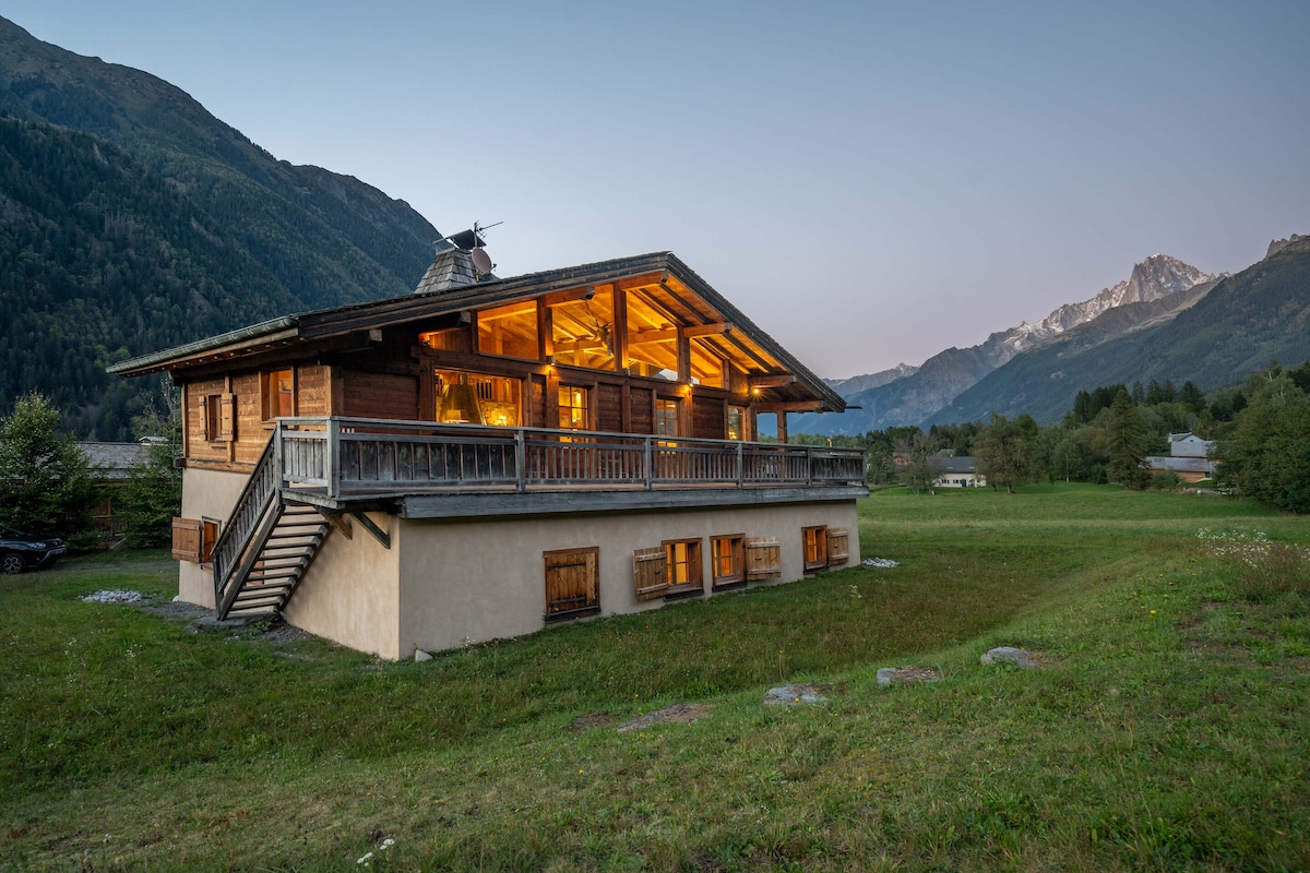 The chalet's exterior is highlighted by a spacious balcony that offers mountain views, framed by large windows. The traditional wooden architecture complements the surrounding landscape, while the softly lit interior suggests a welcoming ambiance. The exterior layout is surrounded by grassy areas and mountain views.