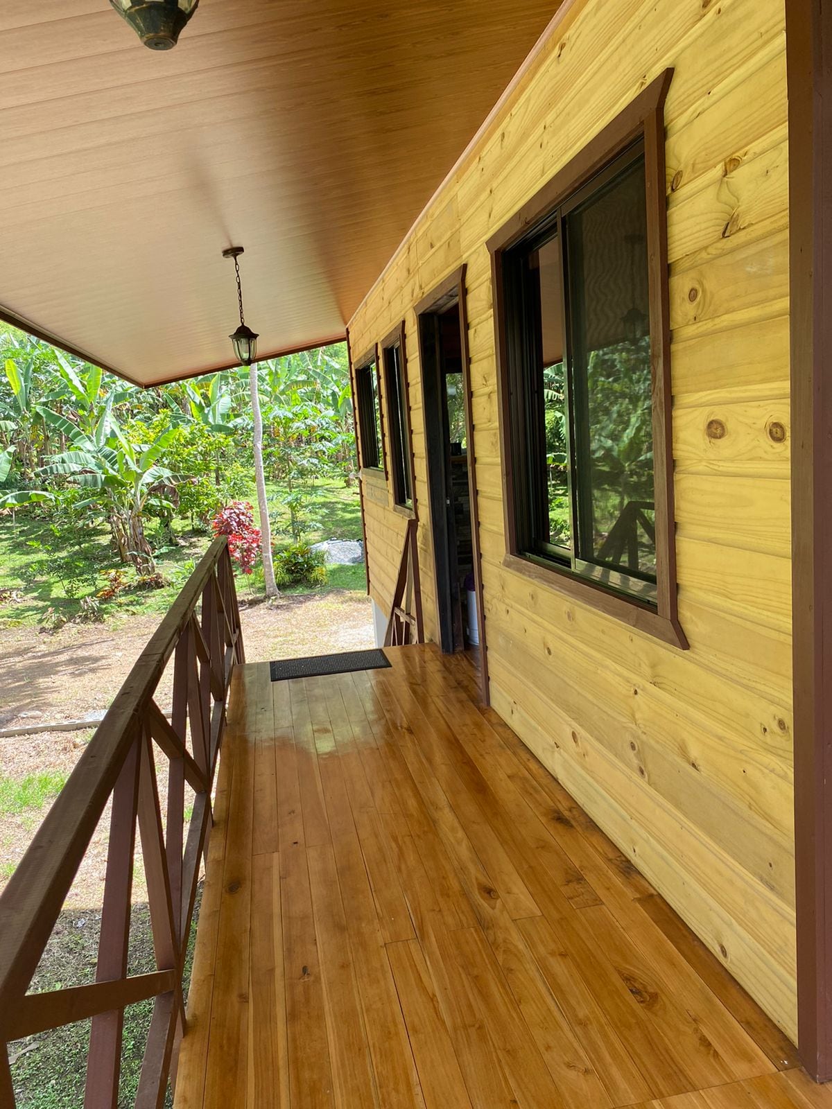 A wooden deck extends along the exterior of the villa, featuring a smooth, polished floor. Large windows are framed in brown, providing views of the surrounding greenery. Lush plants and trees can be seen in the background, emphasizing the natural setting.