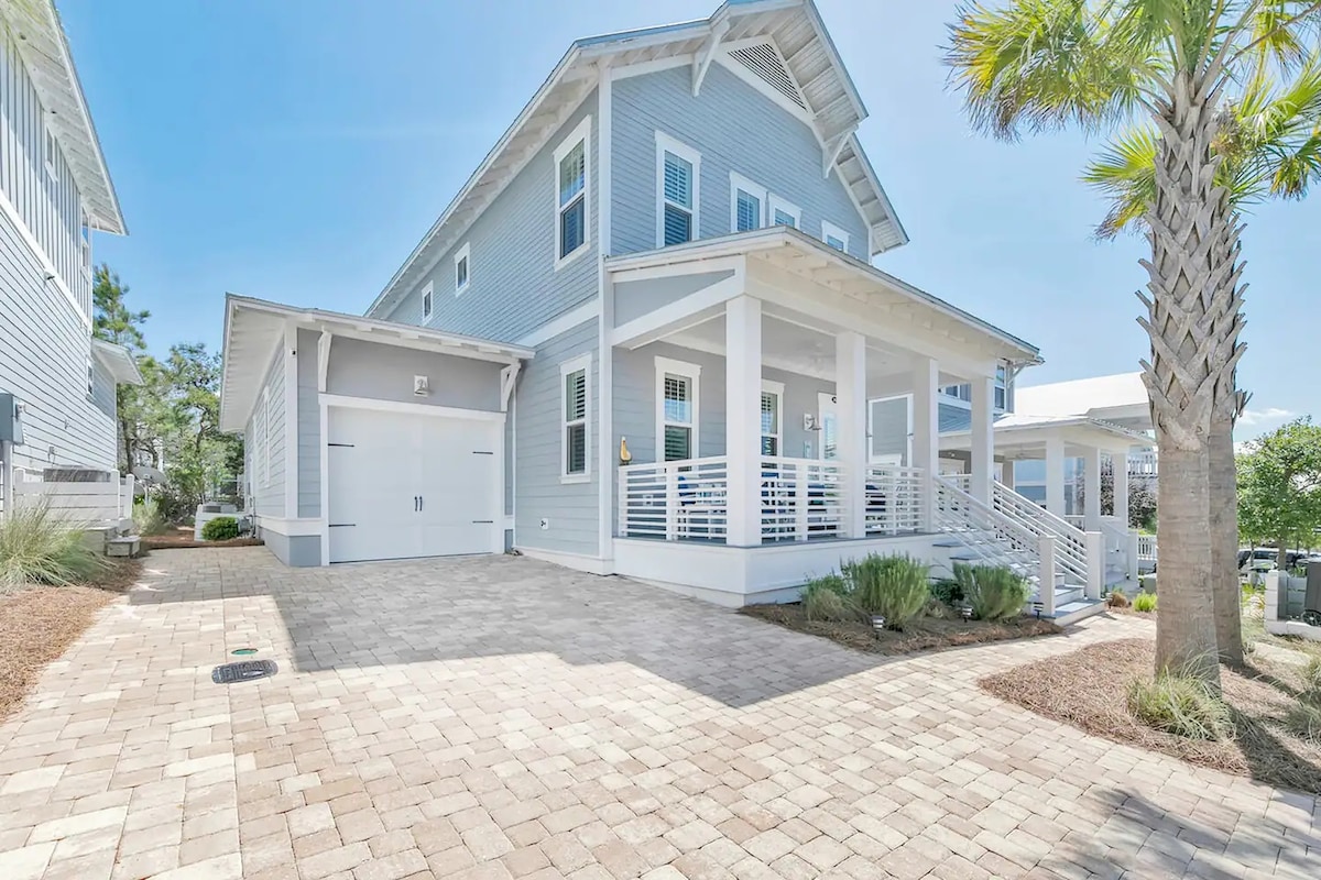 A modern two-story house is shown with a light blue exterior and a spacious paved driveway. A covered porch with white railings is visible at the entrance, surrounded by well-maintained landscaping. The clear blue sky presents a sunny atmosphere.