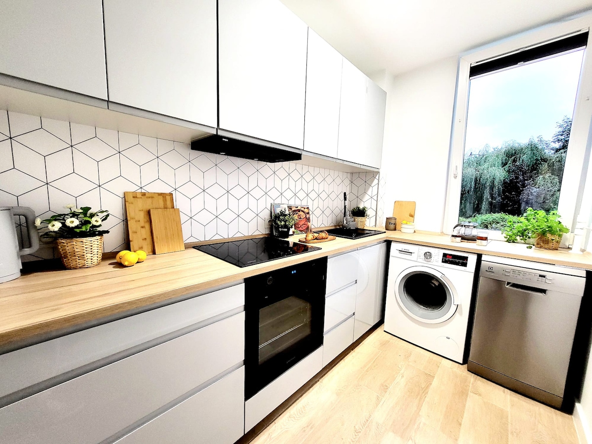 A modern kitchen features sleek cabinetry and a geometric tile backsplash. A wooden countertop provides workspace, with appliances like a dishwasher and washing machine neatly integrated. Fresh herbs and flowers are displayed near a window that offers views of greenery outside.
