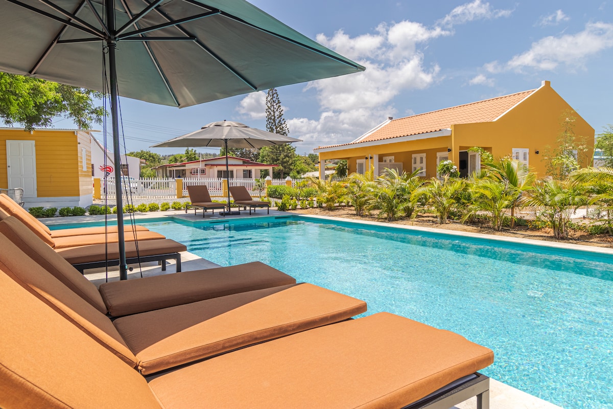 Loungers with dark cushions are arranged by a swimming pool, surrounded by well-maintained greenery. Umbrellas provide shade over the loungers, while a bright yellow building is visible in the background, framed by blue skies and scattered clouds.