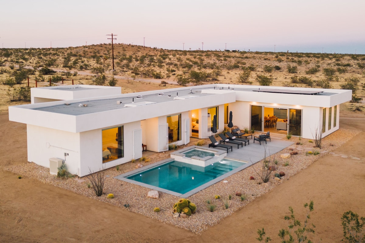 An aerial view of a modern architectural home set against the expansive desert landscape. The property features a saltwater pool and spa, surrounded by well-maintained landscaping. Large windows offer clear views into the interior, where outdoor seating is arranged for relaxation.