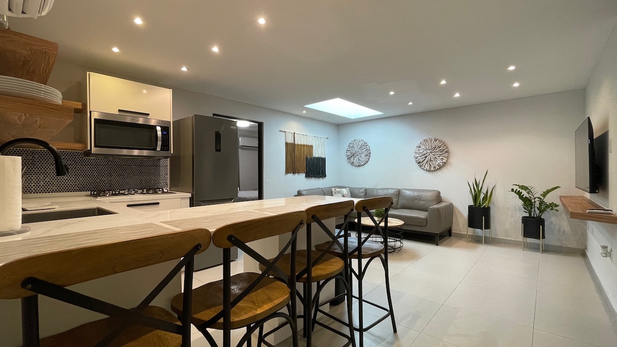 An open-concept kitchen and living area is displayed, featuring a light wood dining table with high-backed chairs. A sleek kitchen includes modern appliances and a stylish backsplash, while a comfortable sofa is set against the wall, complemented by decorative plants and soft lighting.