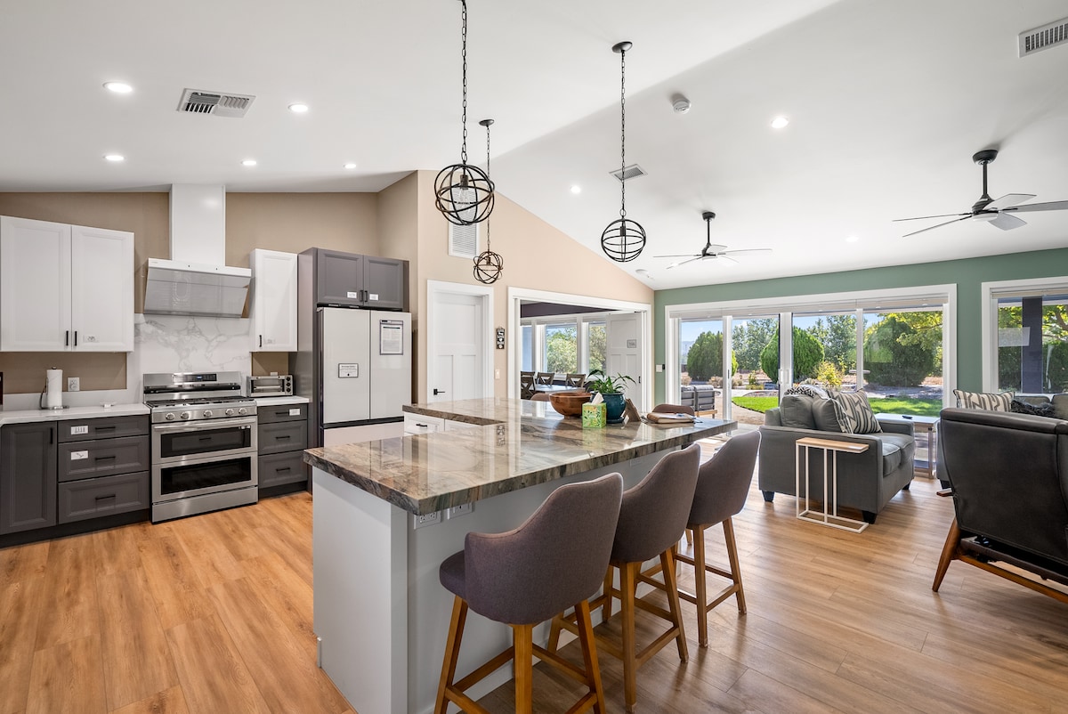 A modern kitchen features a large island with seating for three, complemented by stylish pendant lighting. Stainless steel appliances, including an oven and refrigerator, are arranged neatly. The open layout connects to a cozy living area, visible through expansive windows that offer views of the backyard.