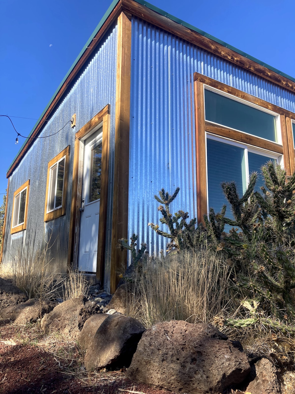 The exterior of the blue metal structure is framed by natural landscaping, featuring native vegetation and rocky soil. Large windows allow natural light to enter the space. The entrance door is visible, complemented by rustic wood detailing, creating a harmonious connection with the surrounding environment.