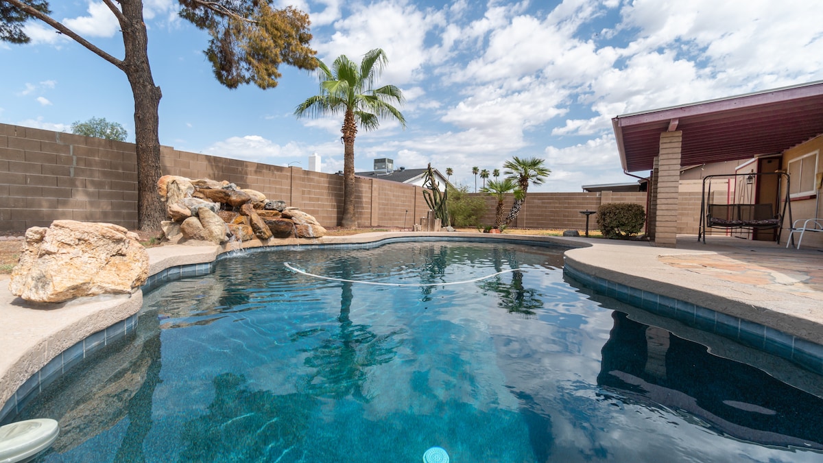 A private backyard features a sparkling diving pool with clear water surrounded by an extended patio. Towering trees and various desert plants enhance the natural setting, while a rocky feature adds visual interest against the smooth pool edge.