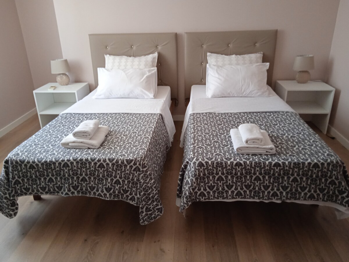 The room features two single beds with soft white bedding and patterned gray throws. Each bed is flanked by a small bedside table with a lamp. Natural light fills the space, highlighting the light-colored walls and wooden floor.