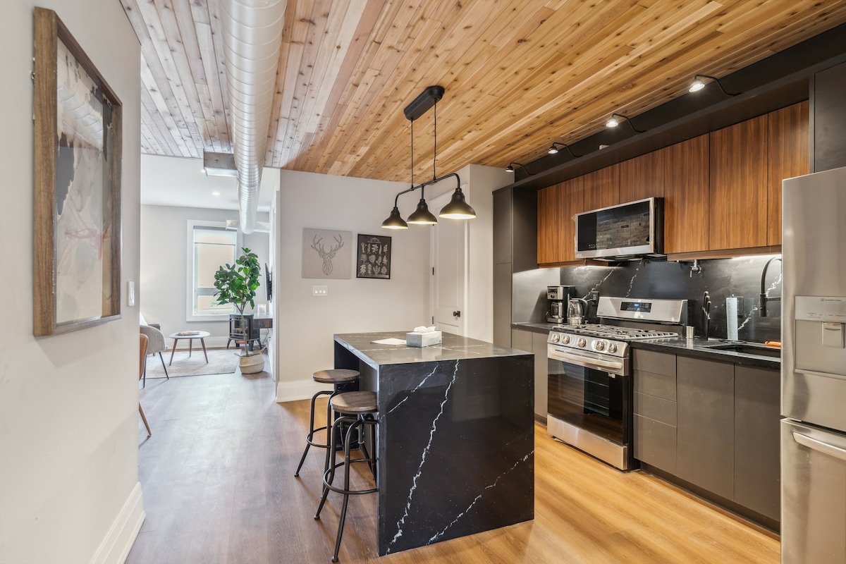 The kitchen features modern stainless steel appliances, including a gas stove and oven. A stylish black island with seating creates a functional space. Natural light enters through a nearby window, highlighting the wood-paneled ceiling and contemporary decor.