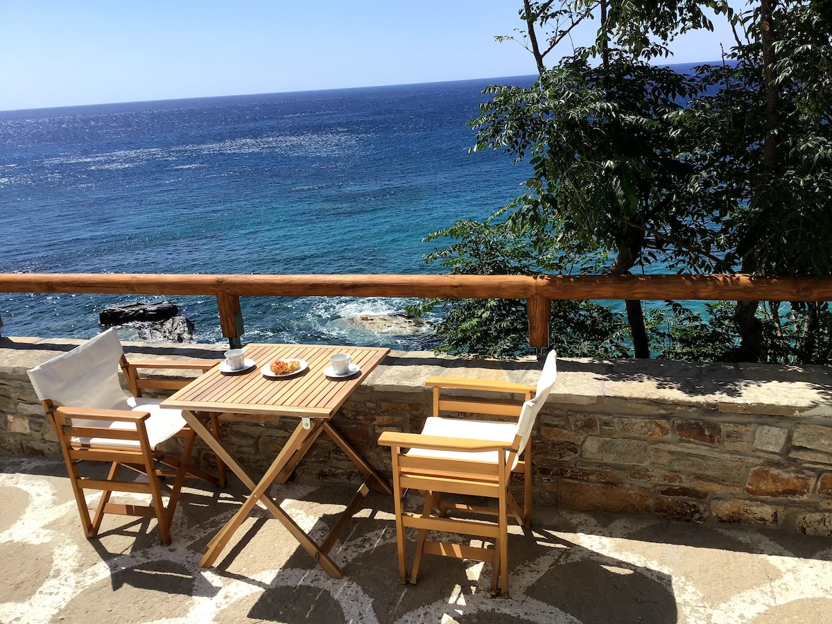 A small wooden table with two folding chairs is set on a stone terrace overlooking the Icarian Sea. Plates of pastries and cups are arranged neatly on the table, while the vibrant blue water glimmers in the sunlight, surrounded by lush greenery.