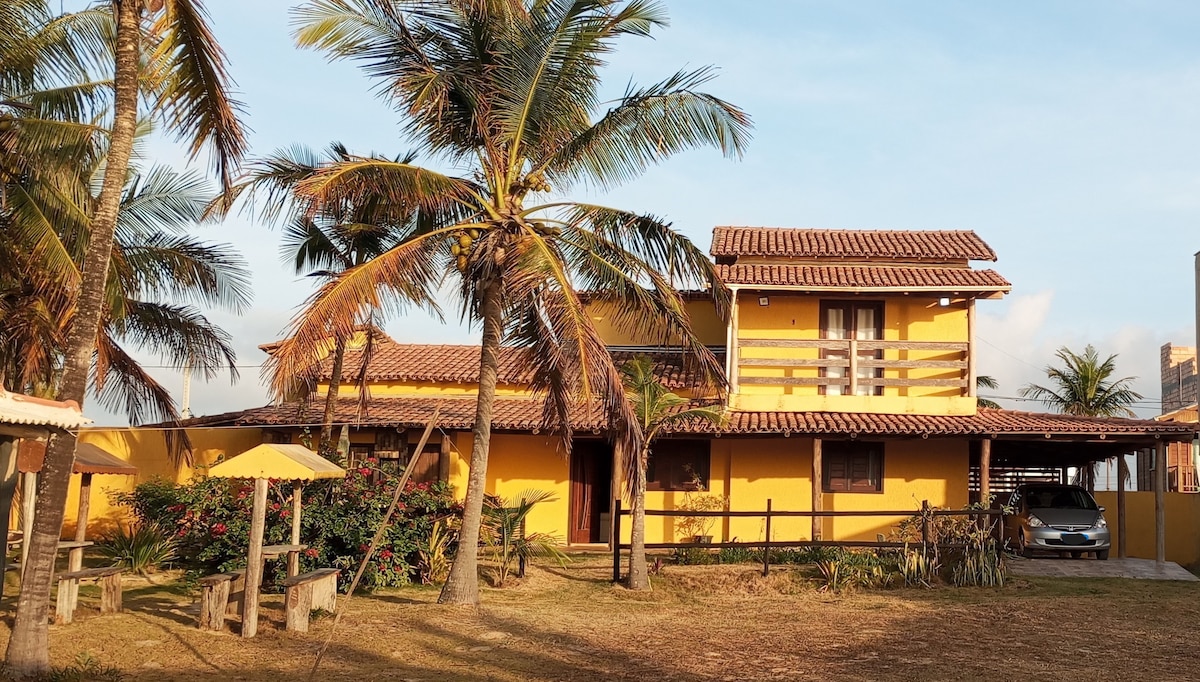 A bright yellow house is set amongst palm trees, showcasing a two-story structure with a tiled roof. Features include layered balconies and large windows. An outdoor area includes a swing set and shaded seating. A car is parked near the entrance, adding to the accessibility.