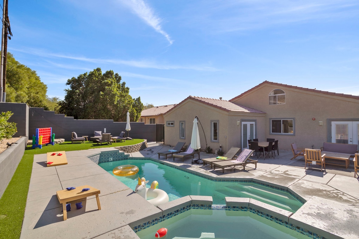 A spacious outdoor area features a wraparound pool with a connected spa, lounge chairs around the pool, and a sunny patch of lawn. Games like cornhole and giant Connect-4 are positioned on the patio. Umbrellas provide shade for seating areas beside the house.