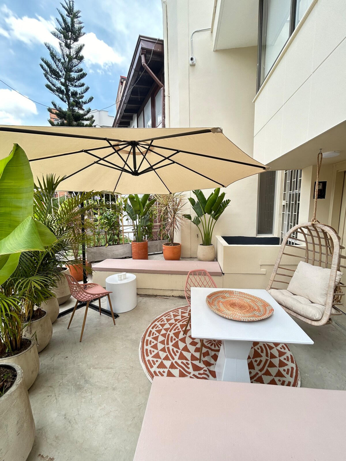 An outdoor patio area is depicted, featuring a swinging seat and a small table adorned with a decorative plate. Potted plants surround the space, and a large umbrella provides shade, creating a calm and inviting atmosphere.