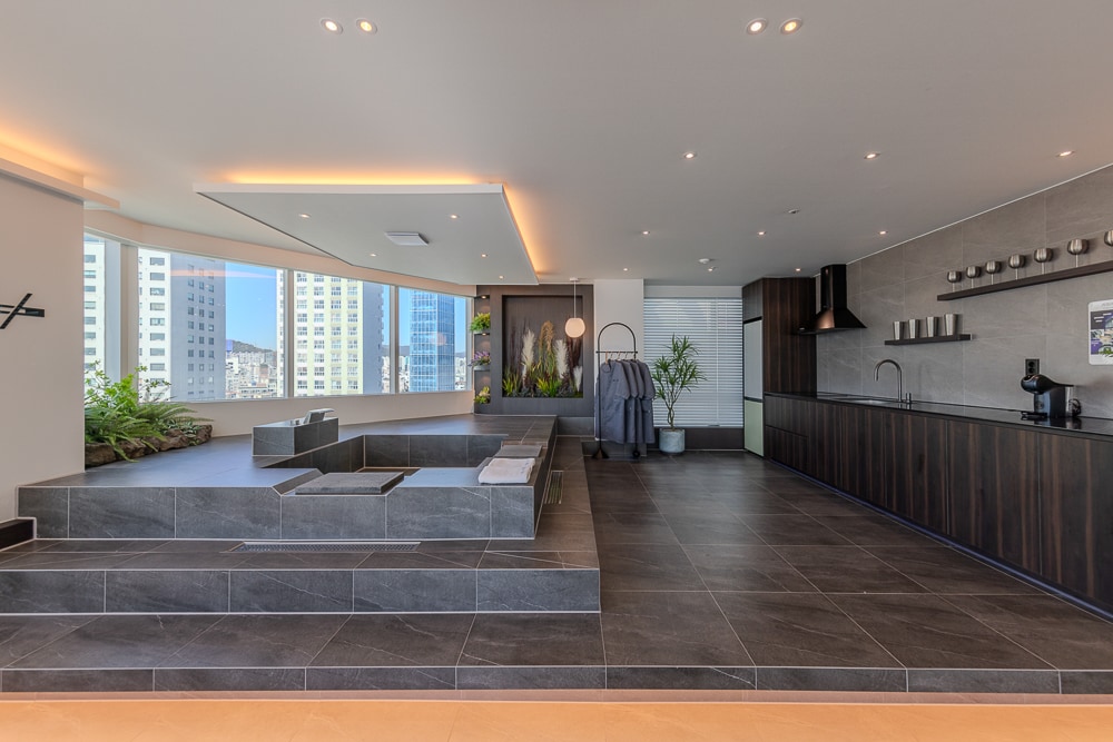 A modern open space features floor-to-ceiling windows, allowing natural light to fill the room. A sleek kitchen is fitted with dark cabinetry, while an elevated seating area with soft seating creates a cozy gathering spot. Greenery adds a vibrant touch to the decor.