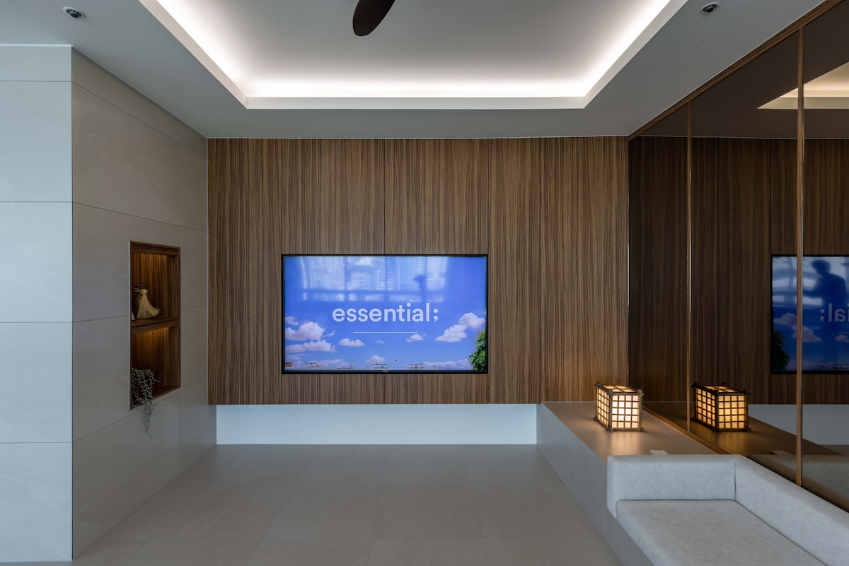An elegantly designed living area features a large wall-mounted television displaying serene imagery. The room is accented by warm wooden paneling and ambient lighting, complemented by a modern seating arrangement and decorative lanterns. A reflective surface enhances the spacious feel of the area.