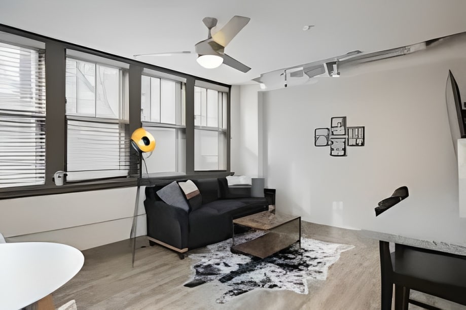 A modern living area features a sleek dark sofa positioned beside a decorative lamp. Large windows bring natural light into the space, accented by stylish artwork on the walls. A minimalist coffee table sits atop a patterned rug, complementing the overall clean design.