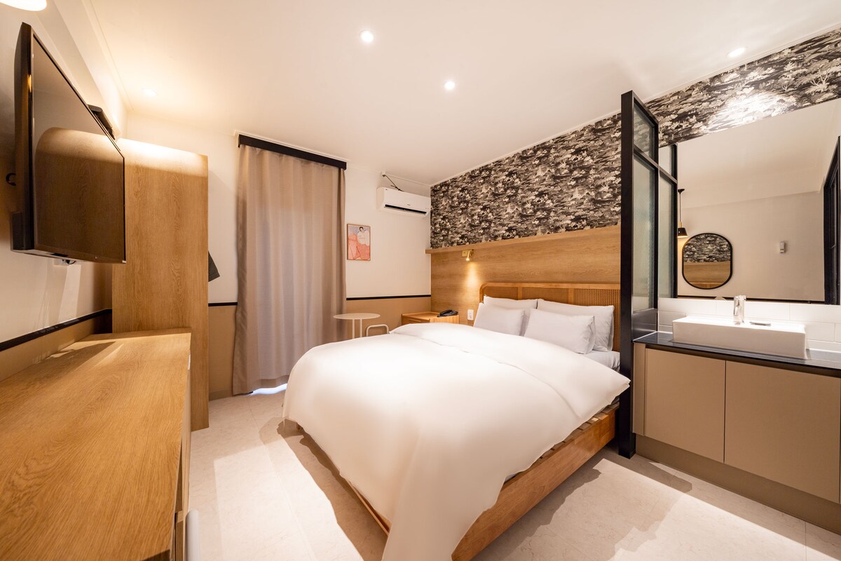 A modern sleeping area featuring a spacious bed dressed in a light duvet. Natural light filters through a partially open curtain, creating a soft ambiance. A stylish wooden dresser and a flat-screen television are positioned against a textured wall, complemented by an elegant wash area.