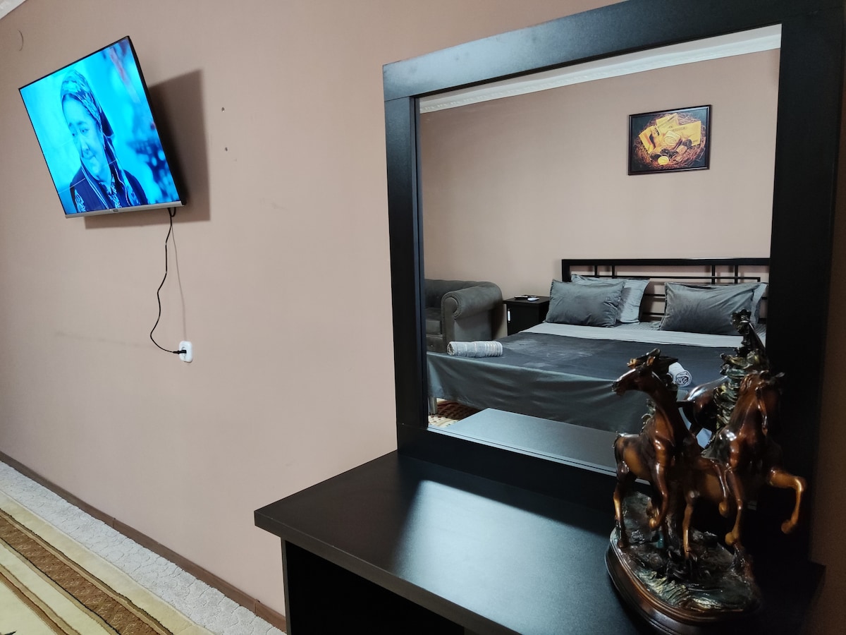A modern room is featured, showcasing a spacious bed with grey bedding. A sleek mirror reflects the room's decor, including a mounted television on the wall. A decorative sculpture of horses is positioned on a dark wood table, adding an element of sophistication.