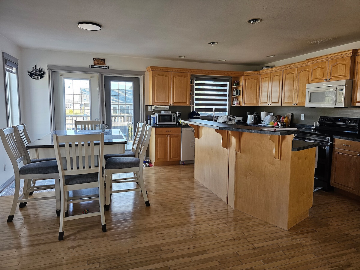 A spacious kitchen area features light wood cabinetry and dark countertops. A dining table with seating for four is situated next to the kitchen island, which offers additional bar seating. Large windows allow natural light to fill the space, enhancing the open layout.