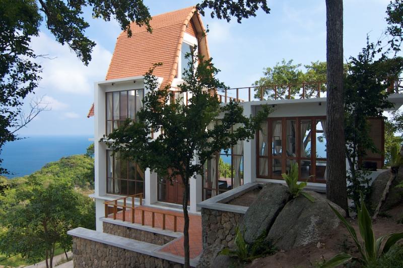 The villa features a unique architectural design with bell-like domes. Large glass windows provide stunning views of the surrounding greenery and ocean. The natural stone and wood elements blend harmoniously into the landscape, creating a serene and inviting exterior.