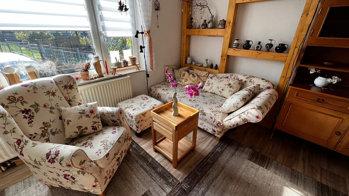 A living area features a pair of floral-patterned armchairs and a matching chaise lounge, arranged around a wooden coffee table. Natural light filters through a large window, illuminating a display of decorative items on a nearby shelf.