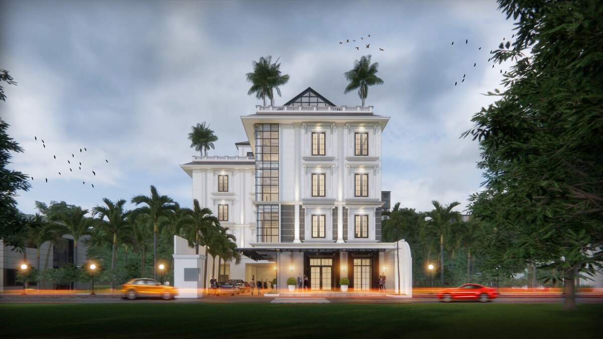 The modern building features a symmetrical façade with large windows and a peaked roof. Flanking palm trees enhance the tropical setting. The entrance is marked by double doors, while soft outdoor lighting creates a welcoming approach to the property.