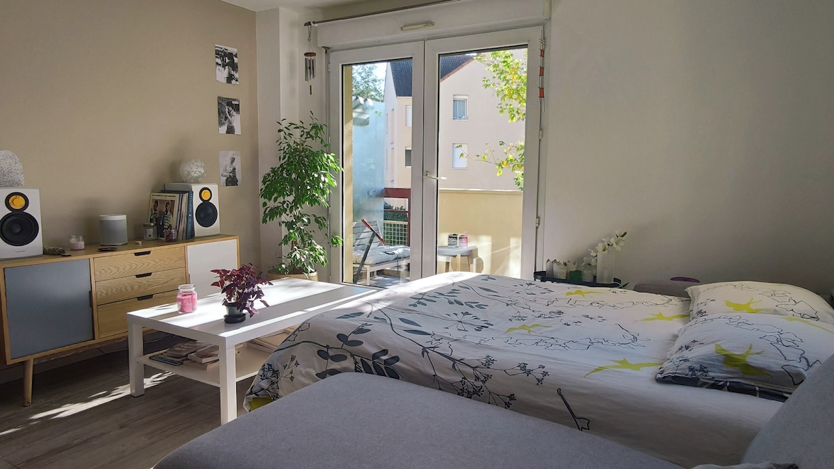 A bright living area features a spacious bed with floral bedding, complemented by a small table and plants. Large glass doors lead to the balcony, allowing natural light to fill the room. Decorative elements include speakers and framed photos on the walls.