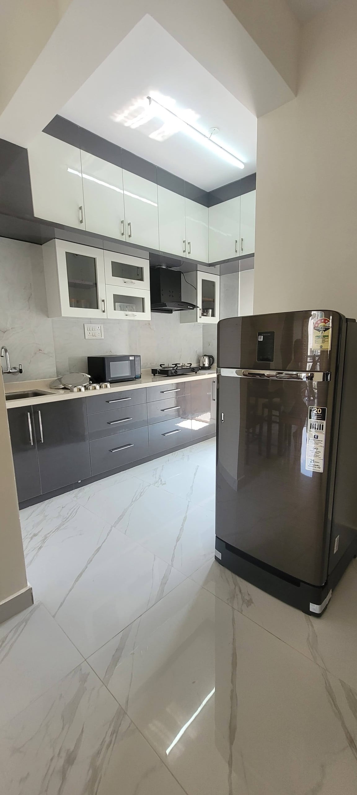A modern kitchen is showcased with sleek grey cabinets and a white countertop. A stainless steel refrigerator stands prominently, reflecting natural light. Cooking appliances, a microwave, and utensils are organized on the countertop, creating an efficient and functional cooking space.