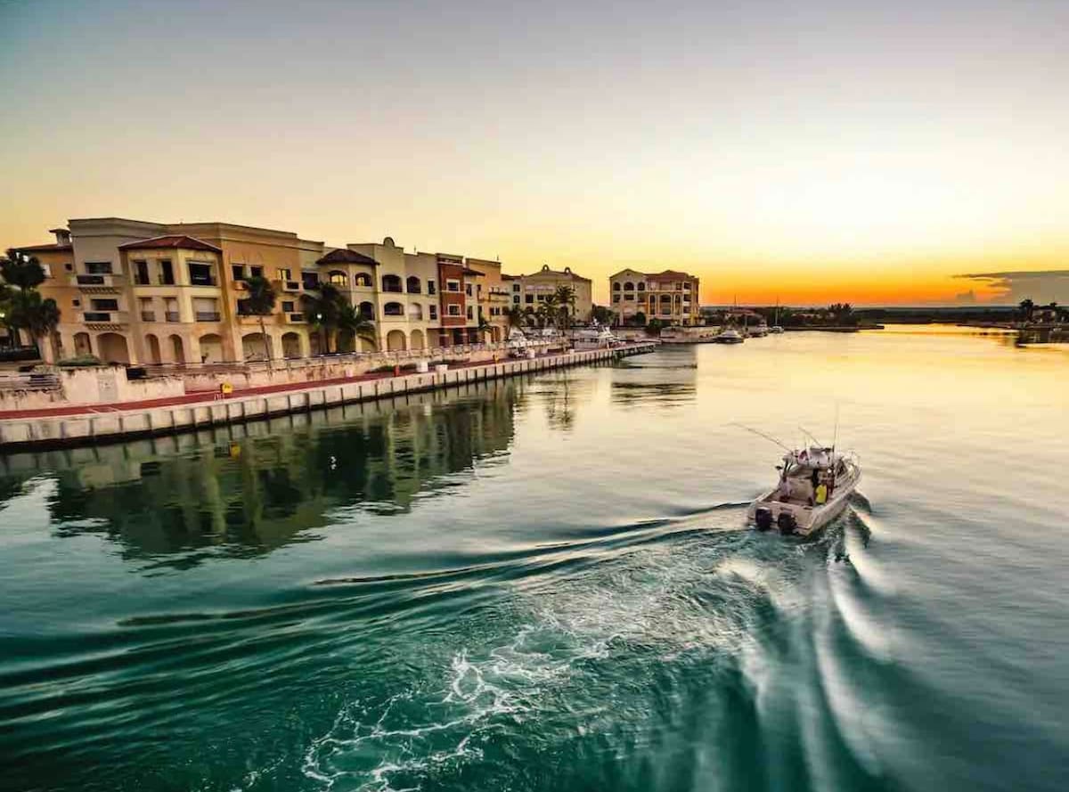 The image captures a scenic view of the marina at sunset, showcasing tranquil waters reflecting the hues of the sky. Elegant buildings line the waterfront, while a boat creates gentle ripples as it moves through the calm water.