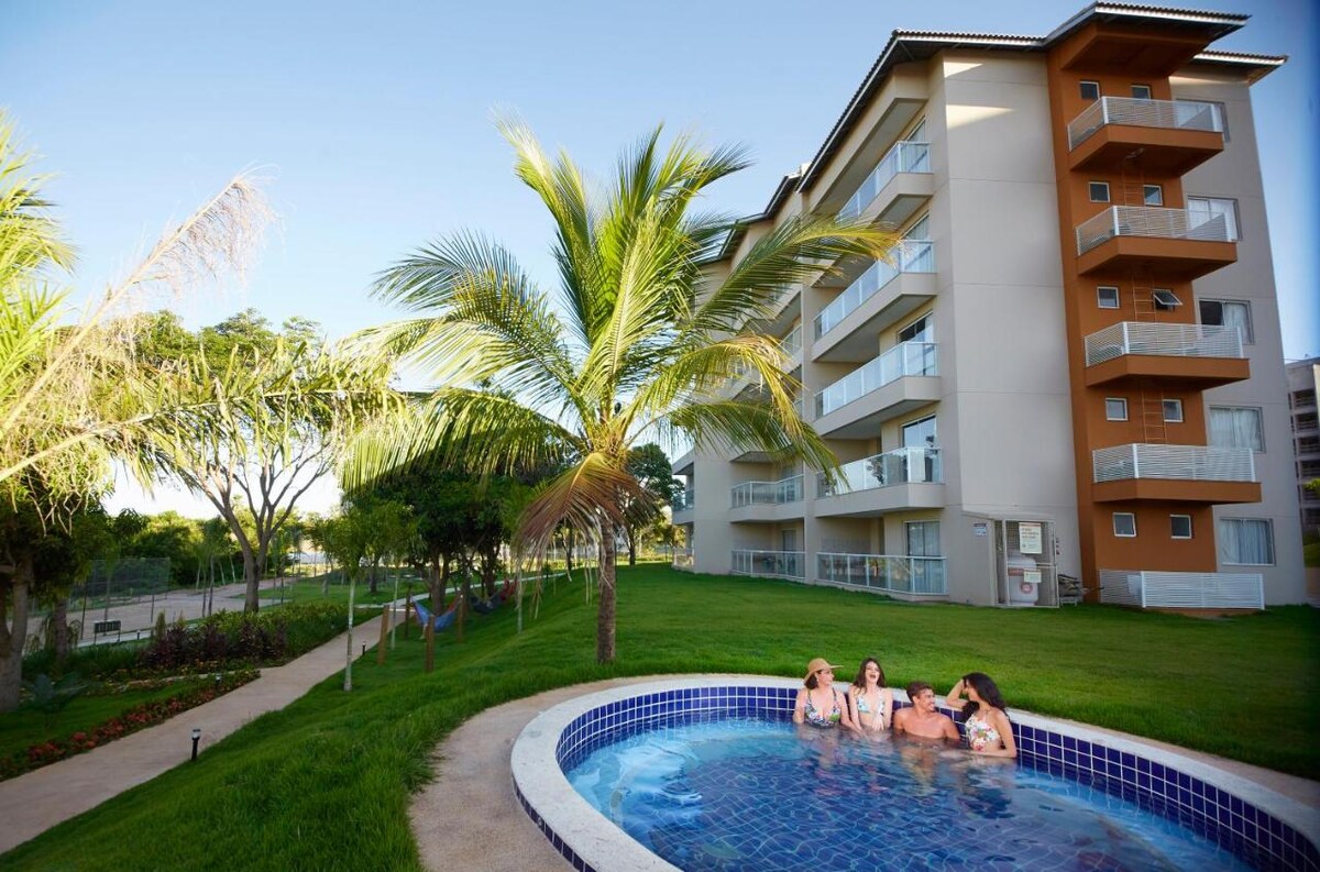 A circular pool is surrounded by a well-maintained grassy area, with palm trees gently swaying in the breeze. A modern three-story building is visible in the background, featuring multiple balconies that overlook the outdoor space.