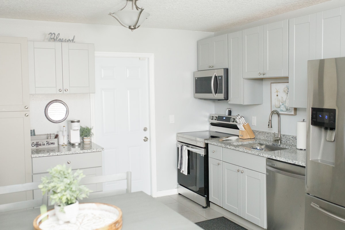 A modern kitchen features light grey cabinetry and a stainless steel refrigerator. A granite countertop is present, complemented by a built-in microwave and stove. Natural light enters through a nearby door, illuminating the space.