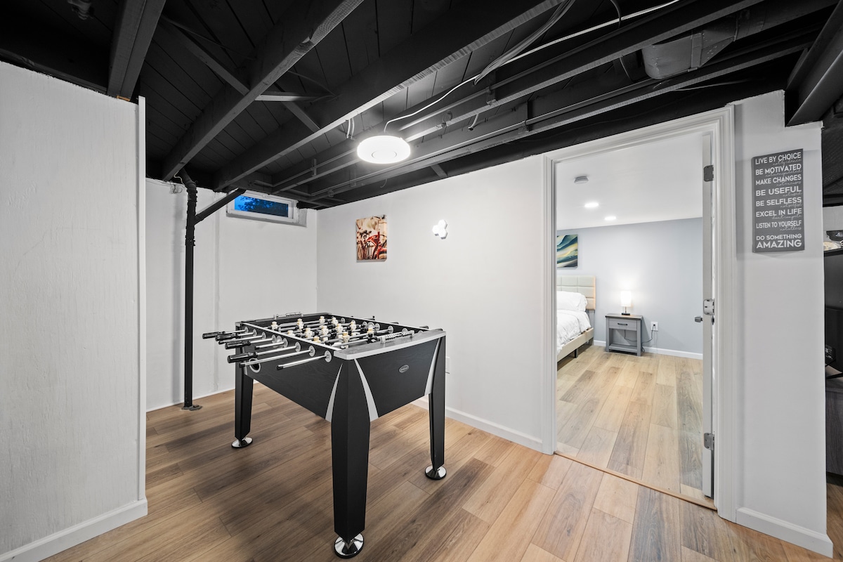 An expansive game room is featured, highlighting a foosball table positioned centrally on a warm-toned hardwood floor. The walls are painted in a light shade, and natural light is allowed in through a nearby window. A doorway leads to another inviting bedroom.