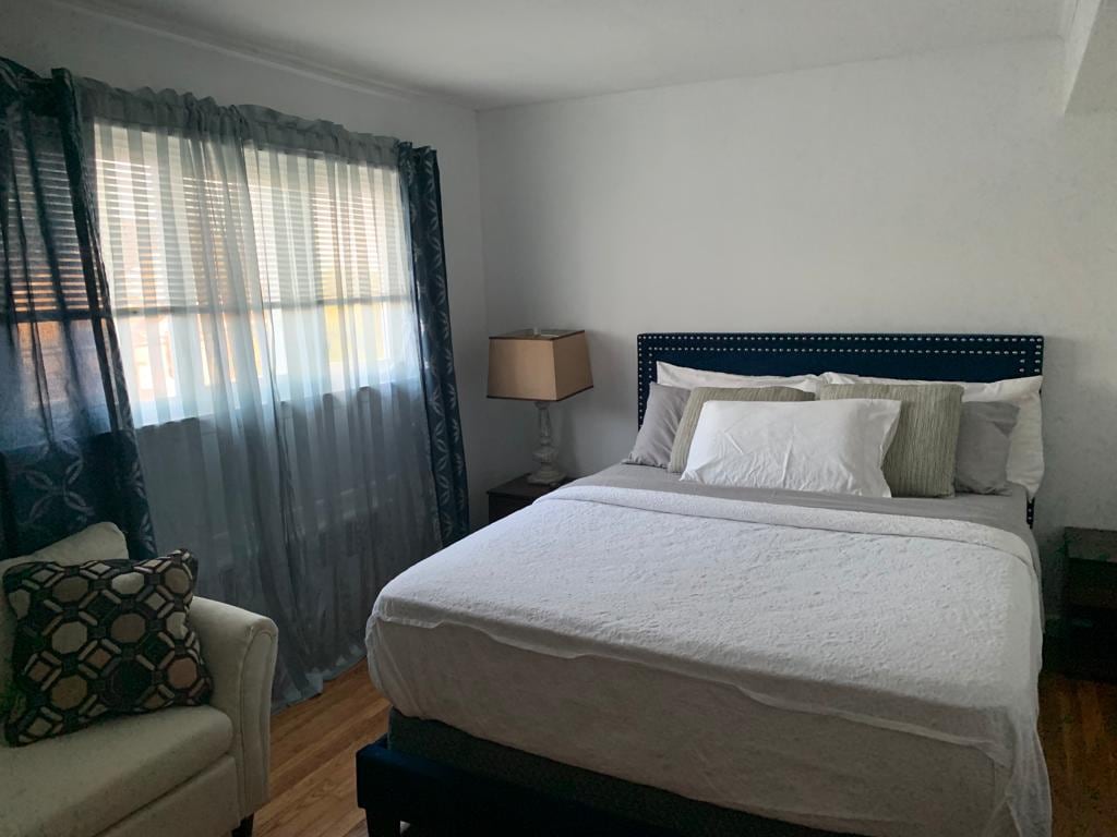 A serene bedroom is featured, showcasing a spacious bed adorned with soft linens and decorative pillows. Natural light filters through a window, complemented by light sheer and dark curtains. A cozy armchair and a lamp provide additional comfort and warmth to the space.