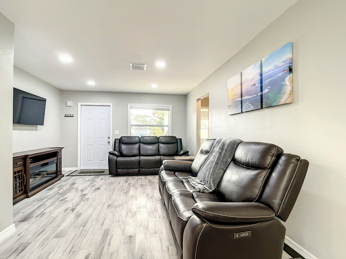 A well-appointed living area features comfortable leather seating arranged for relaxation. A flat-screen TV is mounted on the wall opposite a multi-panel artwork depicting a beach scene. Natural light flows through the window, illuminating the light-colored flooring.