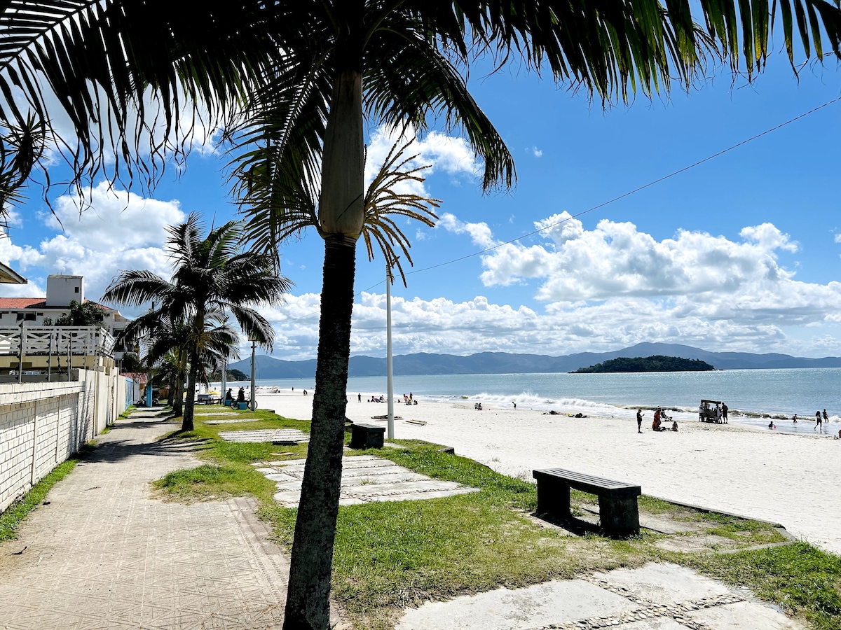 A sandy beach stretches out under a clear blue sky, dotted with visitors enjoying the sun. Palm trees line the path, providing shade along the walkway. In the distance, an island is visible on the horizon, adding to the coastal scenery.