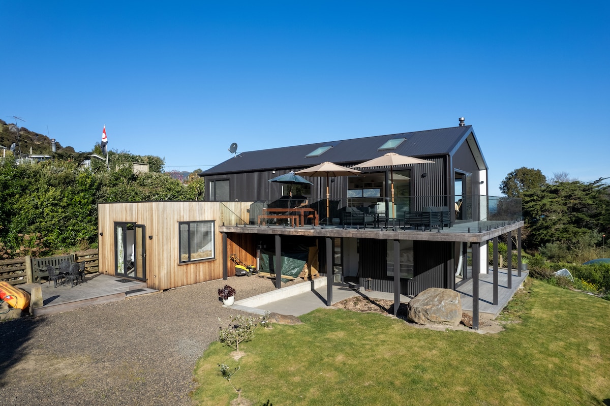 A modern holiday home is set against a clear blue sky, with a spacious deck featuring outdoor seating and an umbrella. The structure combines wood and dark materials, surrounded by well-maintained gardens and rocky landscaping, offering a peaceful atmosphere.