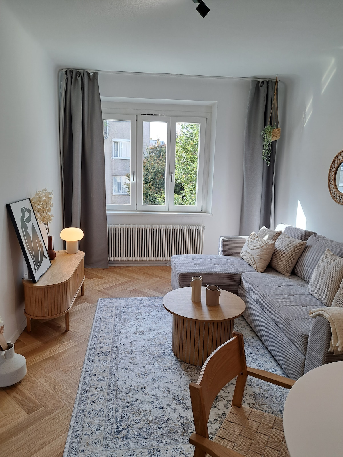 A bright living area features a gray sofa adorned with pillows, positioned next to a round wooden coffee table. A tasteful wall art piece is displayed above a light wooden side table. Natural light enters through a large window, which is flanked by soft curtains.