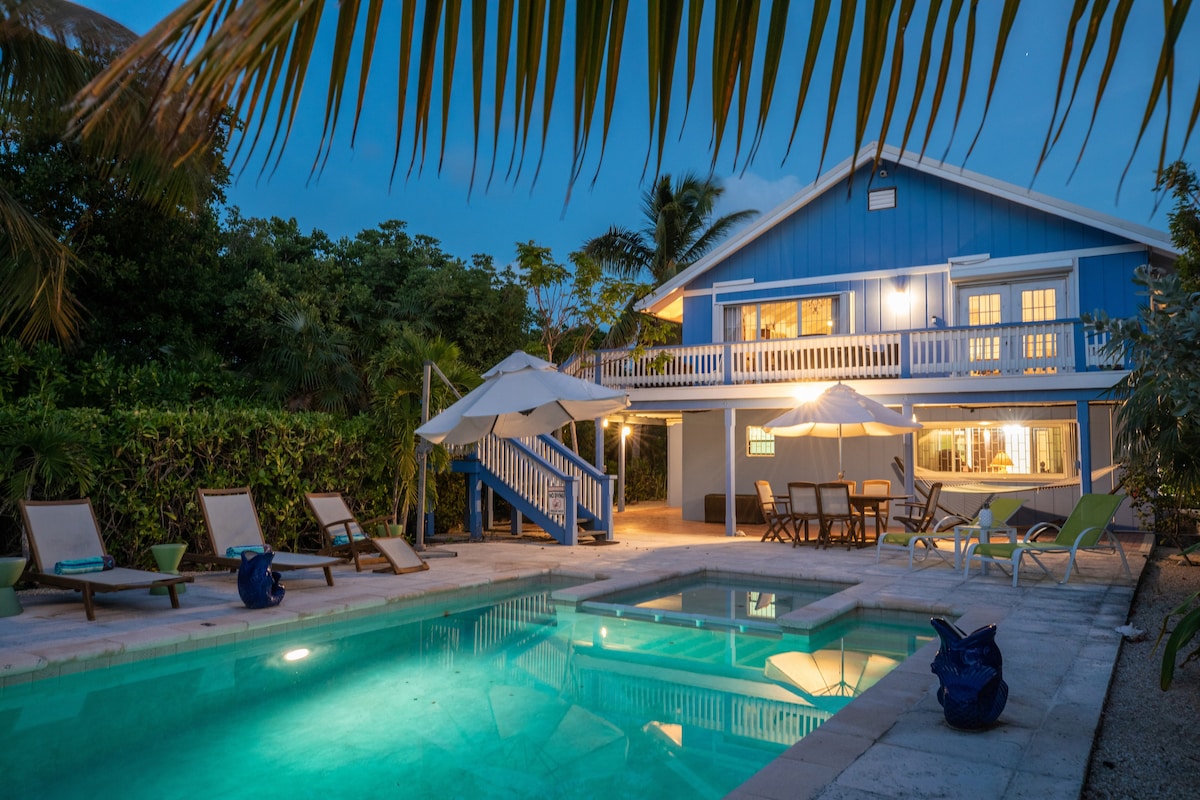 The property is presented in a serene evening setting, featuring a private saltwater pool surrounded by lounge chairs. A spacious outdoor dining area is visible, with an inviting house backdrop highlighted by warm indoor lighting. Lush greenery frames the space, enhancing privacy.