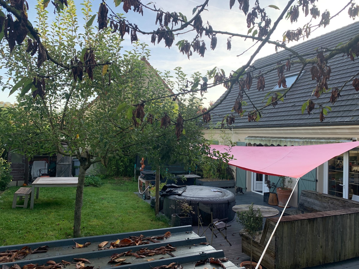 A peaceful outdoor space is depicted, featuring a well-maintained garden with greenery and a few trees. A pink fabric canopy provides shade over a wooden deck area, which includes a hot tub and seating arrangements. Spaces for relaxation and entertainment are visible.