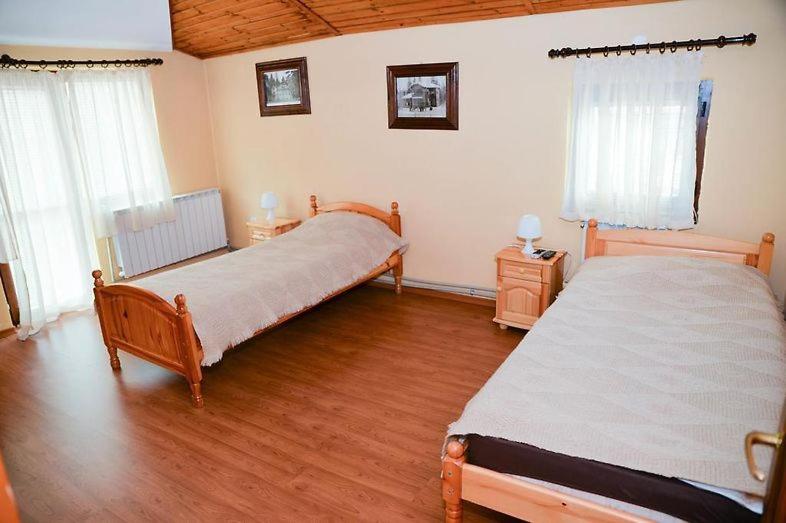 A bright and spacious bedroom features two wooden beds covered with light-colored blankets. Nightstands with lamps are positioned beside each bed. Natural light enters through large windows, softly illuminating the room's warm tones and wooden ceiling.
