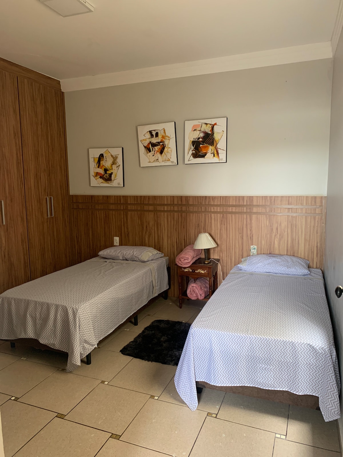 A cozy sleeping area features two neatly arranged beds with soft bedding, complemented by artwork on the wall. A small nightstand with a lamp adds functionality, while textured flooring and wood paneling create a warm environment.