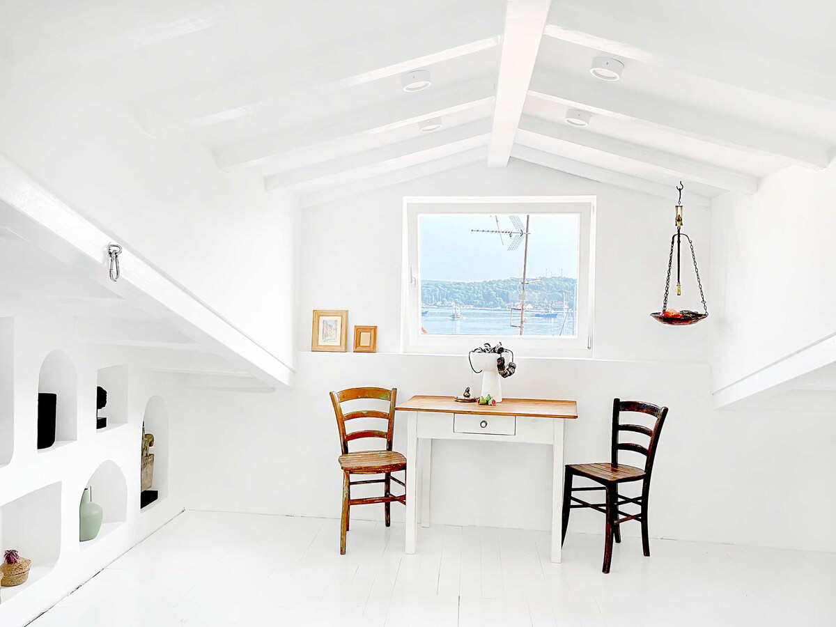 An airy studio space is depicted, featuring a wooden table flanked by two chairs. Bright natural light pours in through a window, offering views of the sea. Decorative elements are subtly arranged on shelves, complementing the room's minimalist white decor.