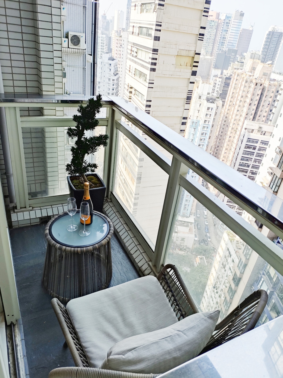 The balcony features a small round table with two glasses and a bottle of champagne set on it. A comfortable chair with soft cushions is positioned beside a potted plant. The view showcases a skyline filled with tall buildings.