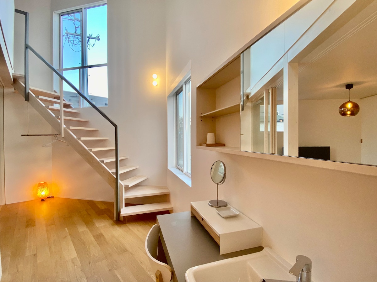 A well-lit interior features a modern staircase leading to an upper level. Natural light enters through a large window, illuminating a minimalist workspace with a mirror and a sink. Warm lighting accents the area, creating a comfortable and functional atmosphere.