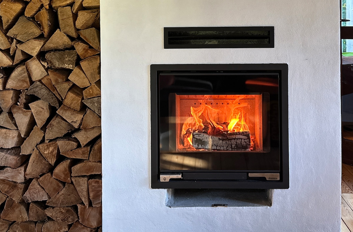 A modern fireplace is displayed with logs burning brightly, surrounded by a neatly stacked pile of firewood. The warm glow from the flames creates a welcoming focal point against the simple white wall.