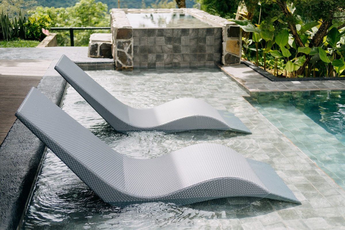 Two modern loungers are positioned partially submerged in a private pool, surrounded by lush greenery and stone accents. A jacuzzi is visible in the background, inviting relaxation and enjoyment of the scenic mountain views.