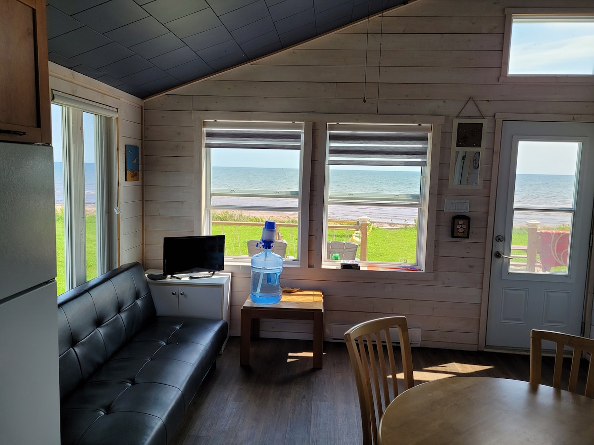 A light-filled interior features a cozy sitting area with a sleek black sofa and a small television. Large windows frame the view of the beach and sea, allowing natural light to fill the space. A wooden table and chairs are positioned nearby.
