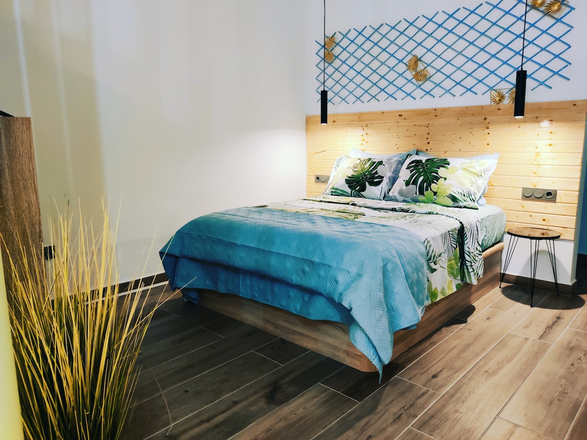 A cozy bedroom featuring a spacious bed dressed in tropical-patterned bedding. A wooden headboard with grid-like accents is complemented by wall-mounted lights. A bedside table stands next to the bed, and decorative grass adds a touch of nature to the modern setting.