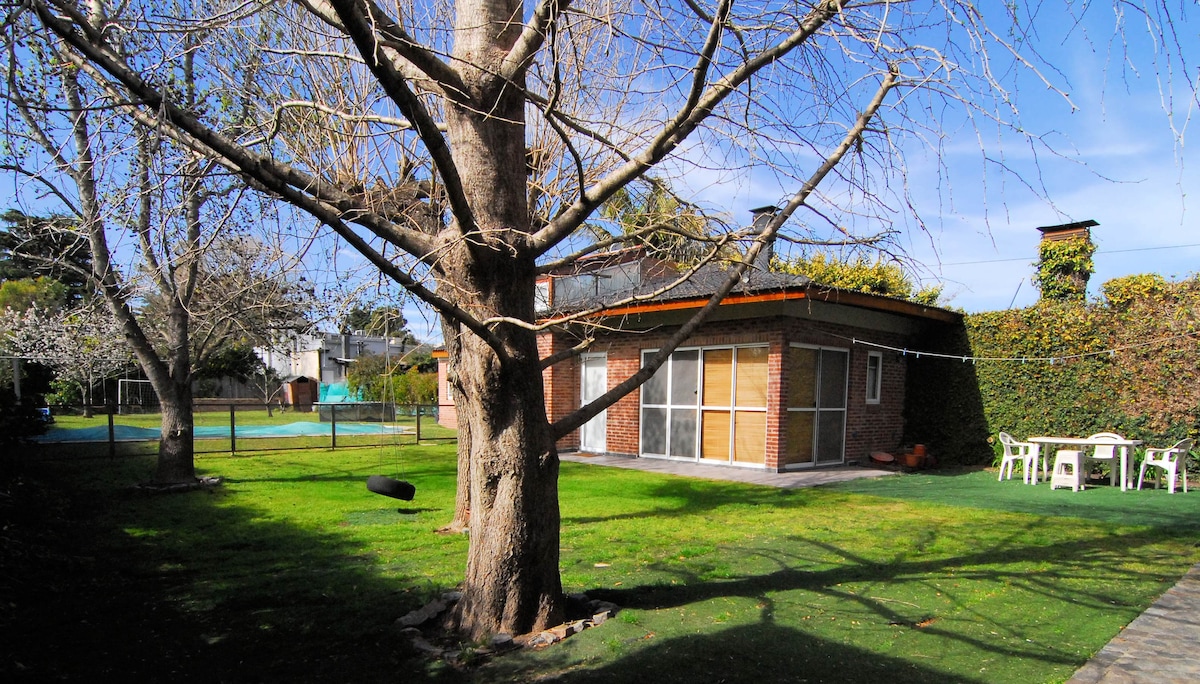 A spacious outdoor area features a well-maintained garden with a large tree and lush green grass. Nearby, a cozy brick structure is accompanied by a seating area with white chairs and a table, positioned against a backdrop of greenery.