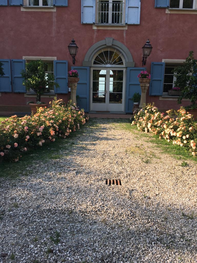 A gravel pathway is lined with blooming rose bushes, leading to a grand entrance featuring large double doors. Flanking the entrance are decorative columns, each topped with potted plants. The building’s exterior showcases soft pink walls and blue shutters.